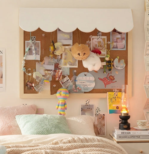 A styled bedroom corner featuring a large scalloped white wall-mounted board covered with photos, notes, clips, and plush toys, above a bed with pastel pillows and a knit blanket.