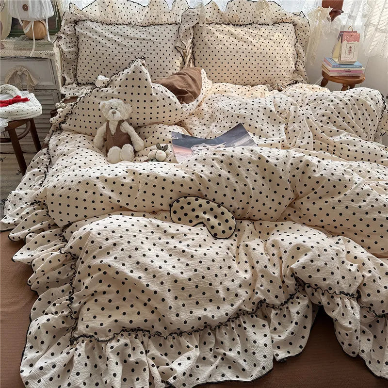 A cozy bed styled with a cream-colored bedding set featuring black polka dots and ruffled edges. The set includes a comforter, matching pillowcases, and a matching eye mask.