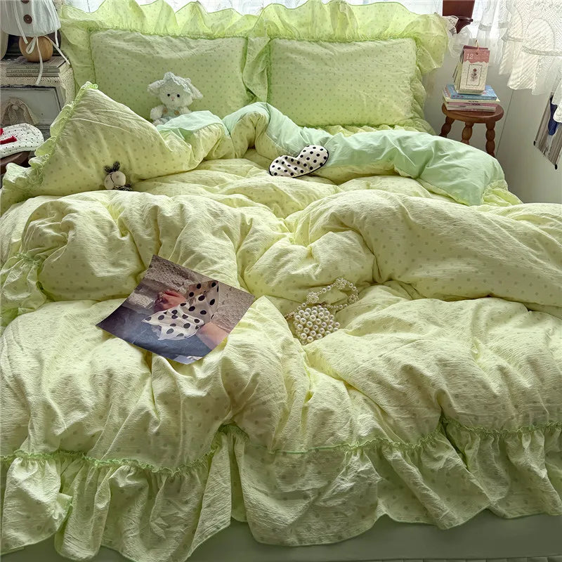 A pastel yellow-green bedding set with ruffled edges and tiny polka dots, styled on a bed with matching pillows, a stuffed sheep, a small plush toy, and a few decorative props in a bedroom setting.