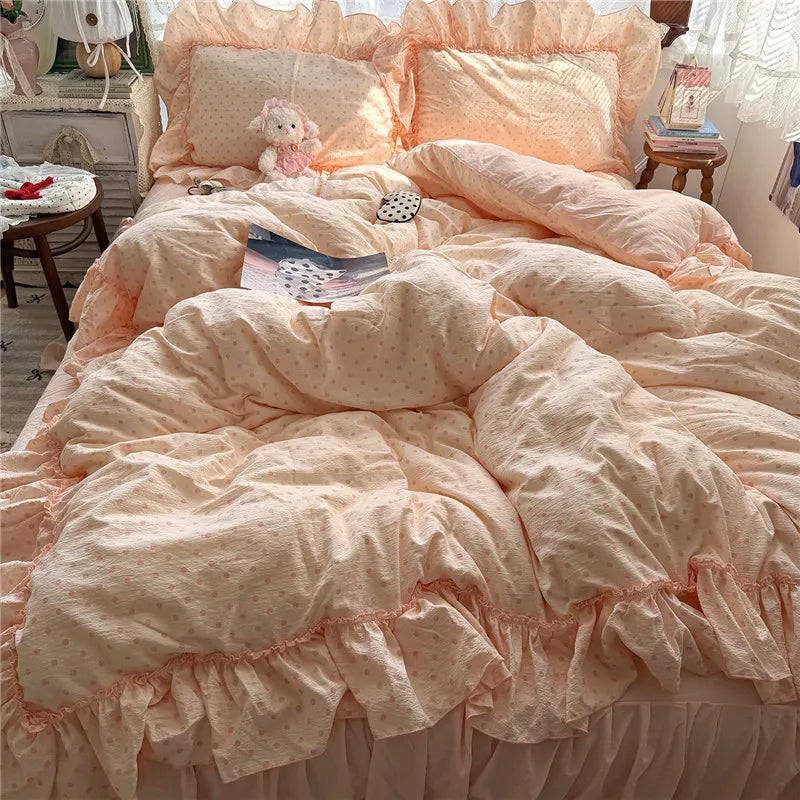 A bed made up with a soft blush pink and white dotted bedding set, featuring ruffled edges, matching pillows, and a ruffled bed skirt in a cozy bedroom setting.