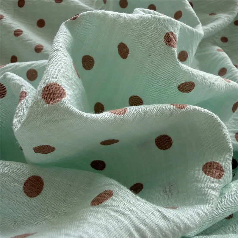 A close-up of a light mint green muslin fabric with a brown polka dot pattern, draped in soft folds to show its texture.