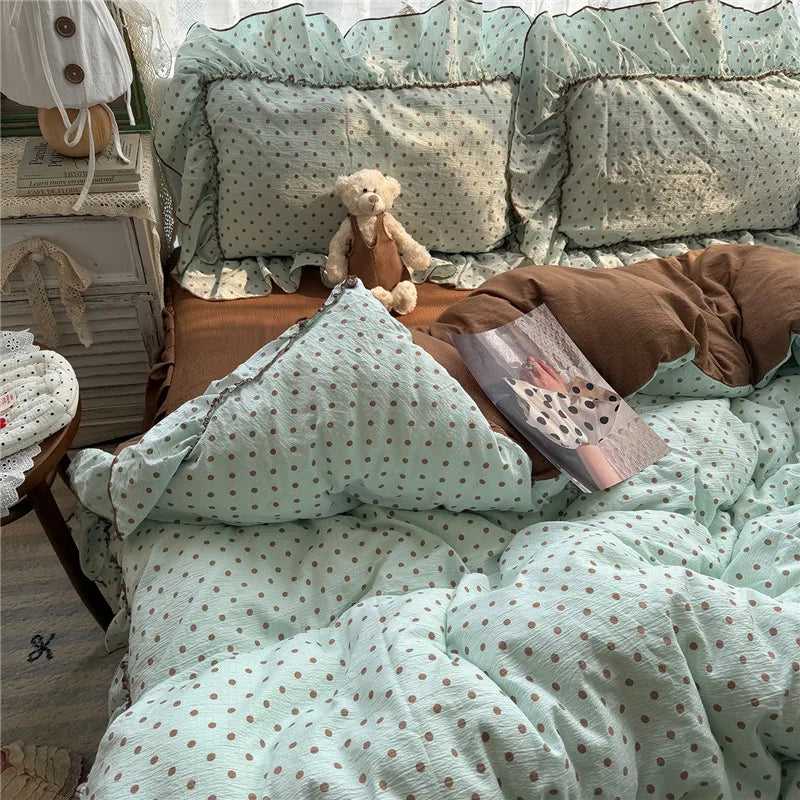 A cozy bed styled with a light mint green and brown polka dot bedding set, including duvet cover, pillows, and a matching pillowcase, with a brown pillow in the center and a teddy bear sitting on the bed.