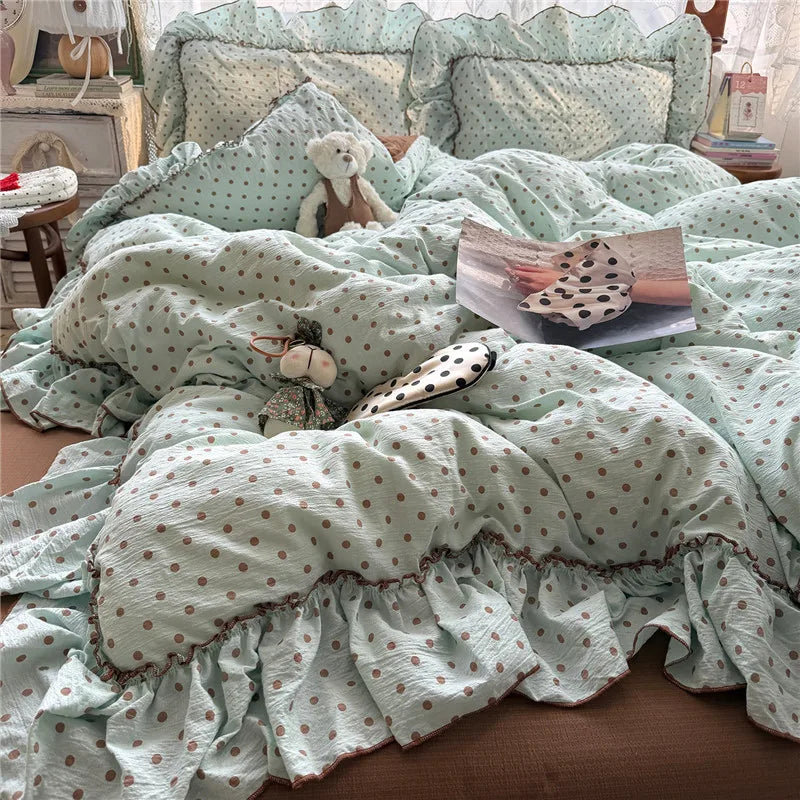 A light mint green bedding set with brown polka dots and ruffled edges, styled on a bed with matching pillows, a teddy bear, and a magazine in a cozy bedroom setting.