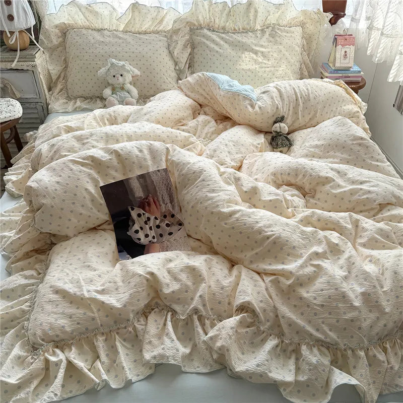A cozy bed made with a cream-colored cotton bedding set featuring tiny polka dots and ruffled edges. The bedding is styled with matching pillows, a small stuffed bunny, and a small plush toy, set in a bedroom with a bedside table and curtains.