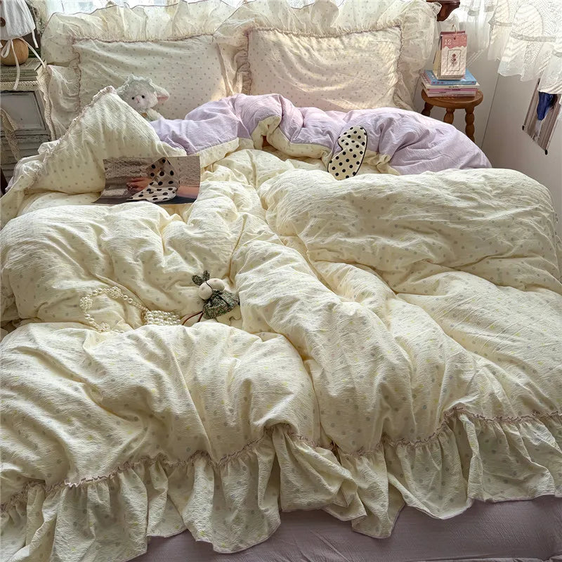 A cozy bed made with a cream-colored cotton bedding set featuring a ruffled hem, dotted pattern, and matching pillowcases, styled with a light purple duvet and decorative pillows in a bedroom setting.