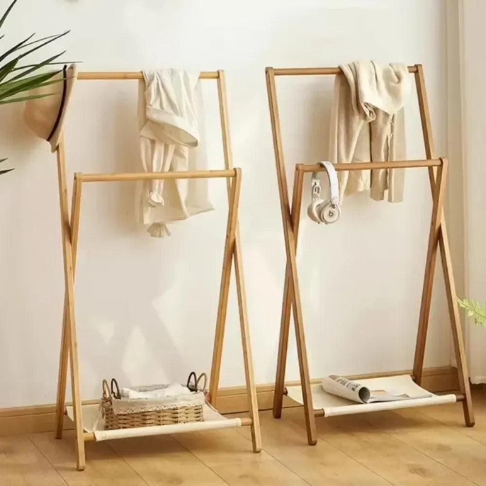 Two light natural wood A-frame clothing racks with white slatted shelves, shown in a bright room with a cream wall and wood floor. One rack has a cream shirt draped on the upper bar, and the other has a cream sweater and white headphones on the upper bar, with a woven basket and magazine on the lower shelf.