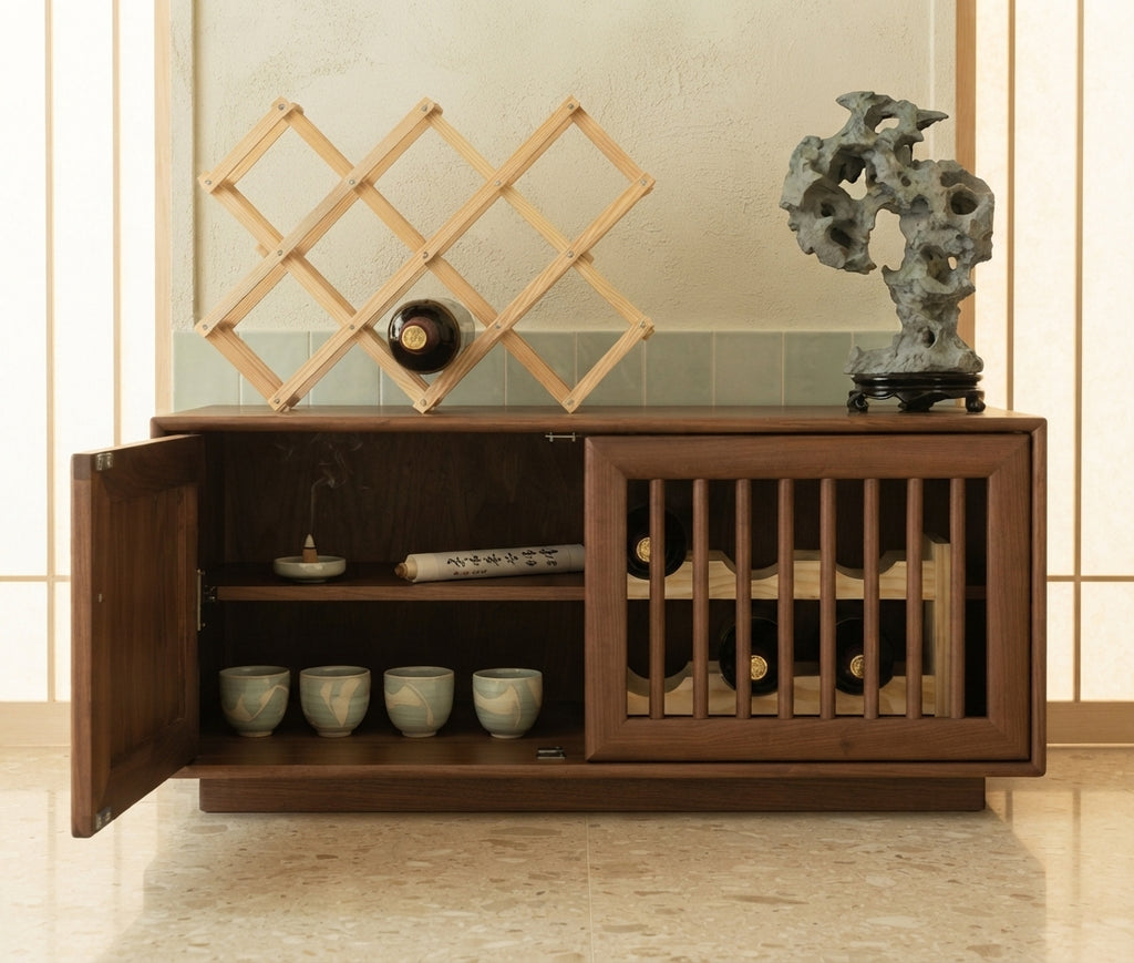 A dark brown wooden wine cabinet with an open left door, showcasing shelves with ceramic cups and a bottle, alongside a slatted right door. A light wood collapsible wine rack and a decorative stone sculpture are placed on top against a light beige wall.